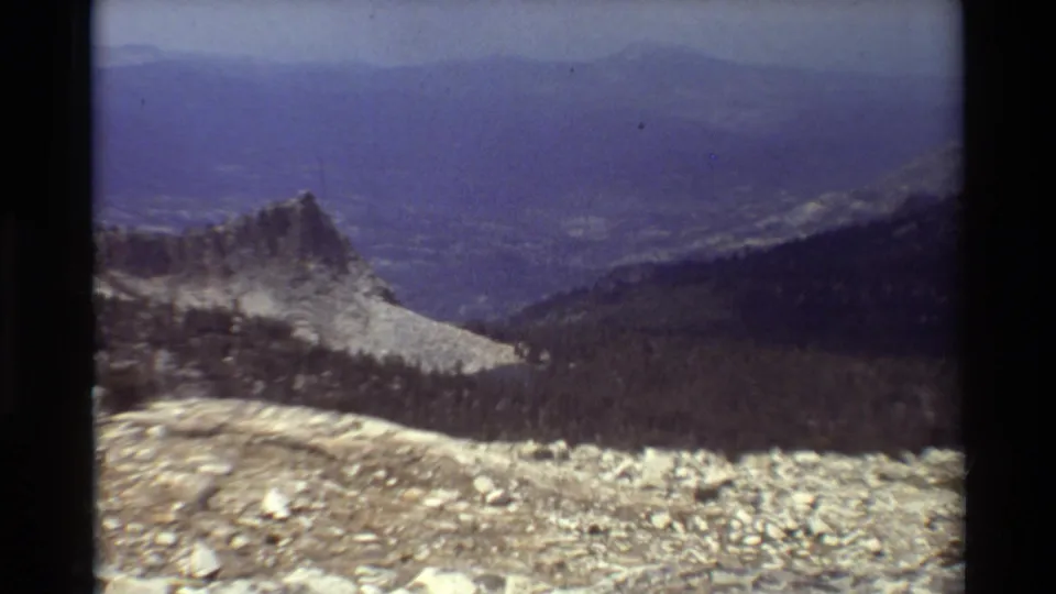 Vintage 8mm frame showing a pale rocky ridge against dark evergreens, with blue atmospheric haze over the distant valley.