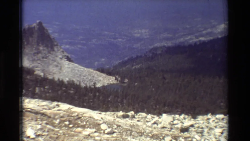 Final 8mm frame of the mountain overlook, with a pointed rocky ridge, tree-covered slopes, distant valley detail, and dark scan borders.