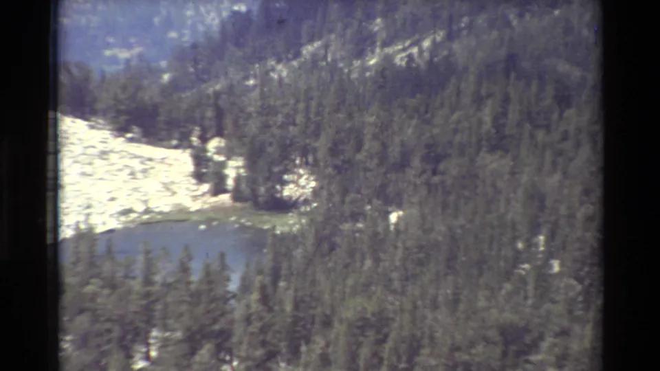 Closer 1981 8mm frame showing a blue alpine pond edged by evergreens, with light rock patches above it.
