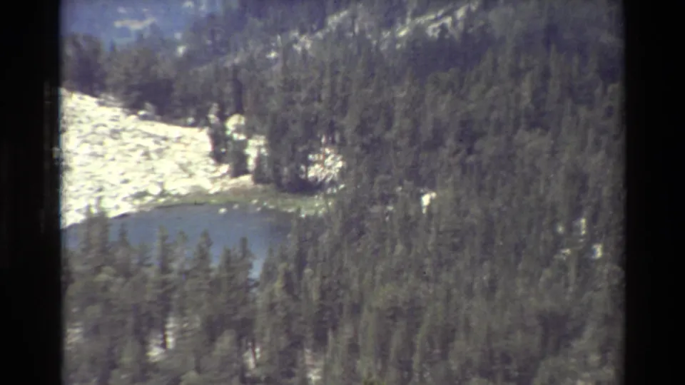 Zoomed 1981 8mm frame of an alpine pond set within dense evergreens below pale rocky mountain slopes.
