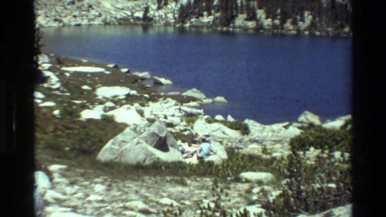 1981 8mm view of a rocky lakeshore in the described British Columbia setting, with two people seated among boulders near blue water.
