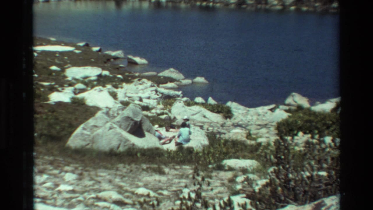 Rocky lake edge with water, boulders, brush, trees, and two seated people becoming clearer.