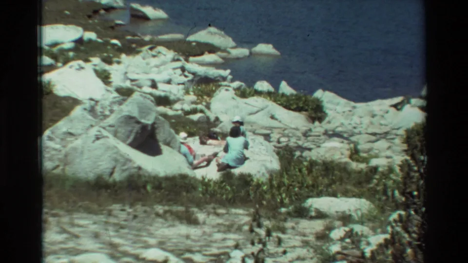Closer 8mm frame showing two people relaxing on shoreline boulders, with blue lake water, pale rocks, and brush around them.