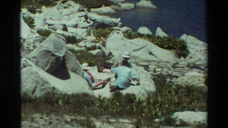 Closest view in the 1981 home-movie clip, showing two people seated on bright shoreline rocks beside a blue lake.