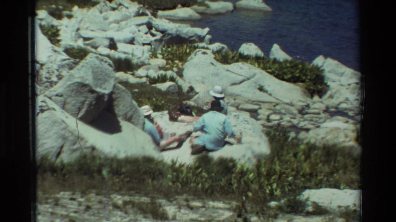 1981 8mm frame of two adults seated on large lakeside rocks in a British Columbia-listed setting, viewed from above.