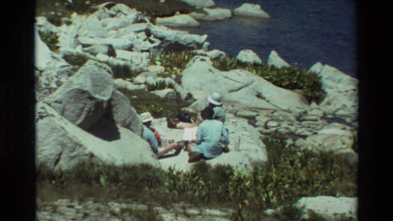 Two men remain seated among boulders above a rocky shoreline.