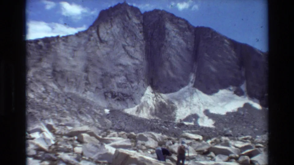 Wide 1981 8mm mountain view in British Columbia, with rocky peaks, snow patches, blue sky, and two distant figures.