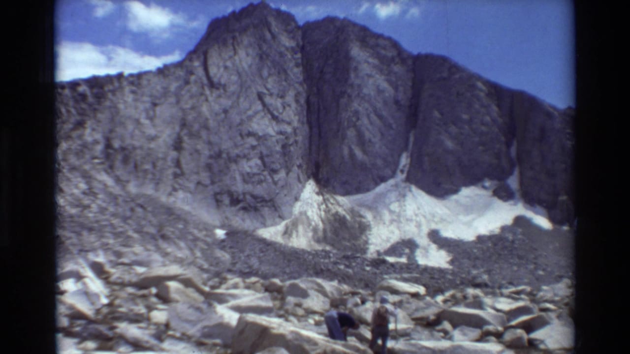 Two distant hikers stand on a rocky slope below a steep gray mountain wall with snow patches.