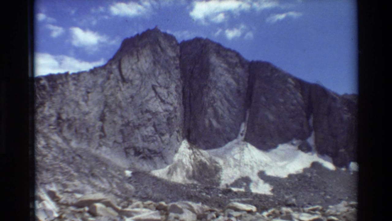 Archival 8mm view of a rugged British Columbia mountain outcrop, snow at the base, and scattered clouds overhead.
