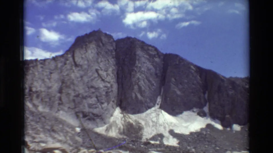 Archival 8mm frame of a steep British Columbia rock outcrop, pale snow patches, and a blue sky with small white clouds.