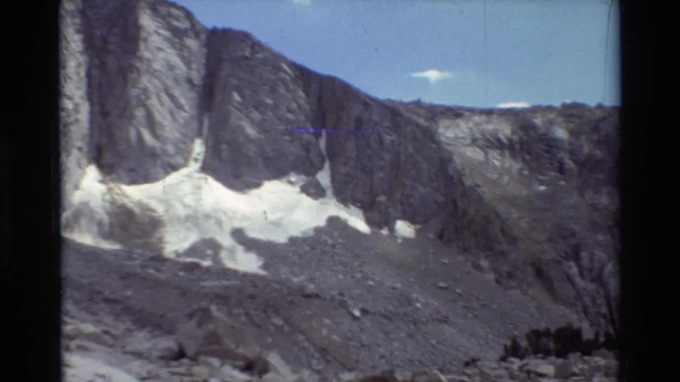 Vintage 8mm frame showing a rugged gray rock face, snow in shaded gullies, and open sky in the British Columbia mountain landscape.