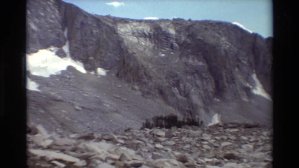 1981 8mm mountain view with a steep rock wall, scattered snow, gray boulder field, and a small dark stand of trees near the lower slope.
