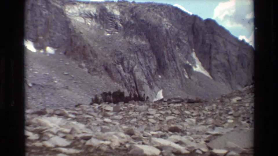 Home-movie frame of a rocky British Columbia mountain slope, showing boulders in the foreground, dark trees, cliff faces, and small snow pockets.