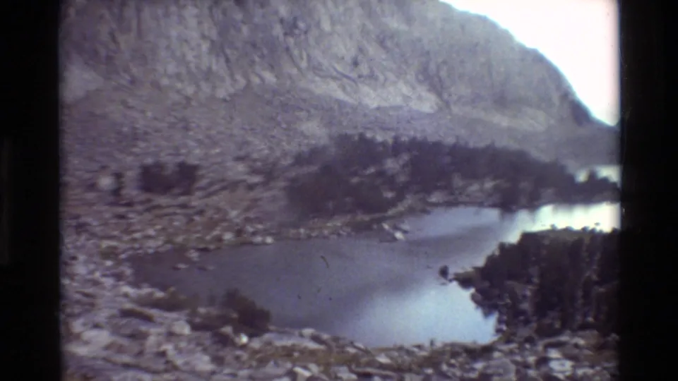1981 8mm frame of an alpine lake below rocky mountain slopes, with evergreen shoreline and calm water visible.