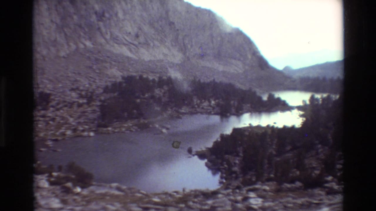 1981 8mm view across an alpine lake toward evergreens, rocky terrain, and a broad mountain face.