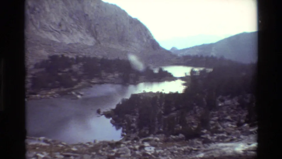 1981 8mm frame showing a closer alpine lake view with trees, rocky shore, and hazy distant ridges.