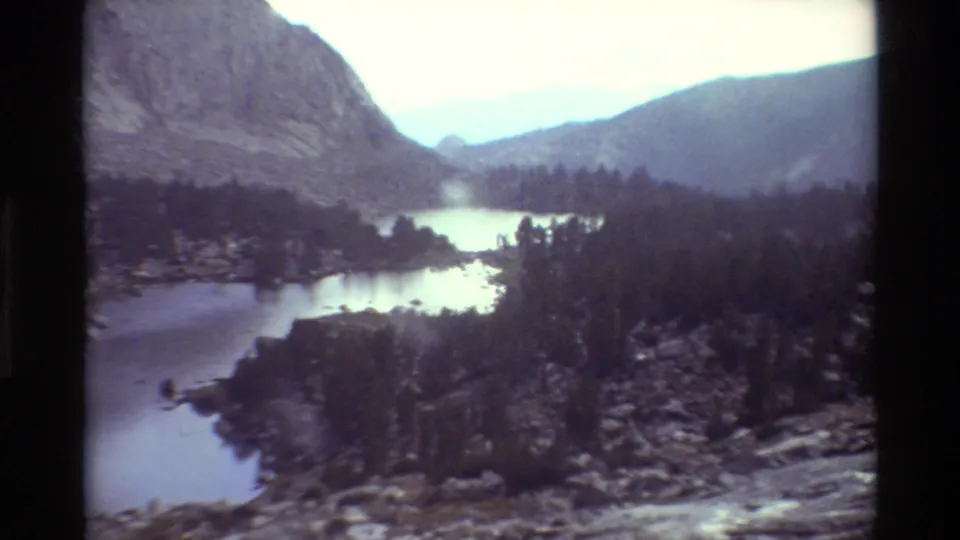 1981 8mm frame of a mountain lake beside dark evergreens, with rocky terrain and hazy ridges in the distance.