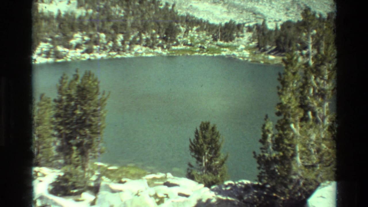 8mm alpine lake view with green-blue water, conifer trees, pale rocks, and a wooded slope, consistent with the supplied British Columbia metadata.