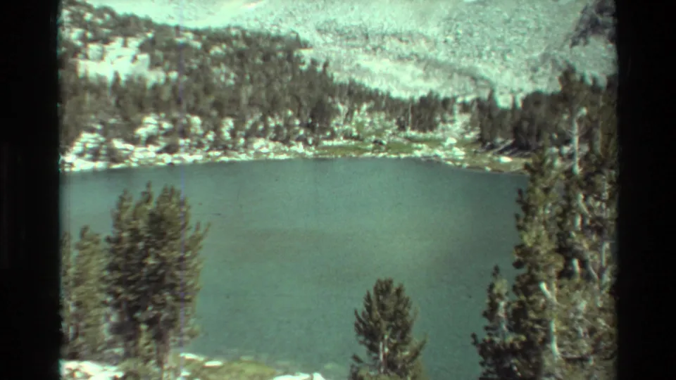 Home-movie frame of a calm mountain lake seen past conifers, with rocky shore and forested hillside under muted 8mm color.