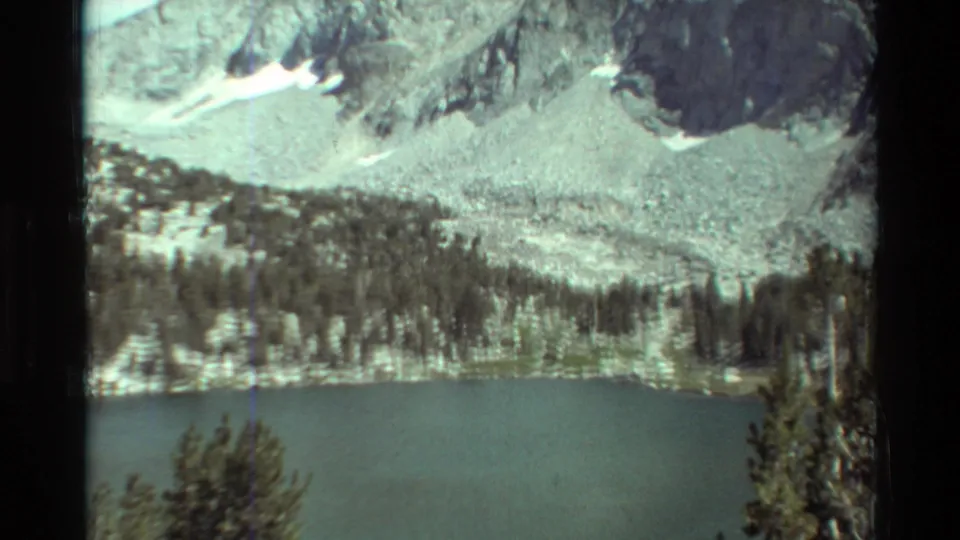 Wide 8mm view of an alpine lake beneath rugged rock faces, scattered snow patches, and a dark evergreen band in the lower slope.