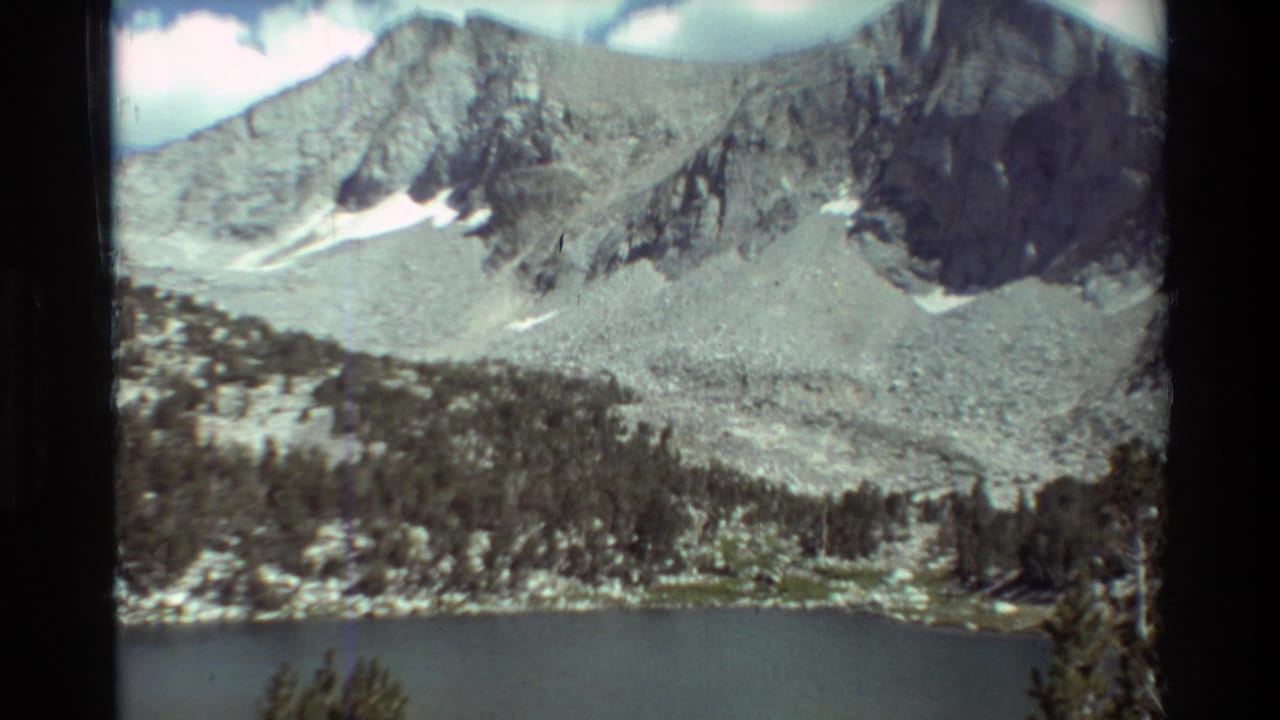 1981 8mm home-movie view of a mountain lake below rugged gray slopes, evergreen trees, clouds, and lingering snow patches.