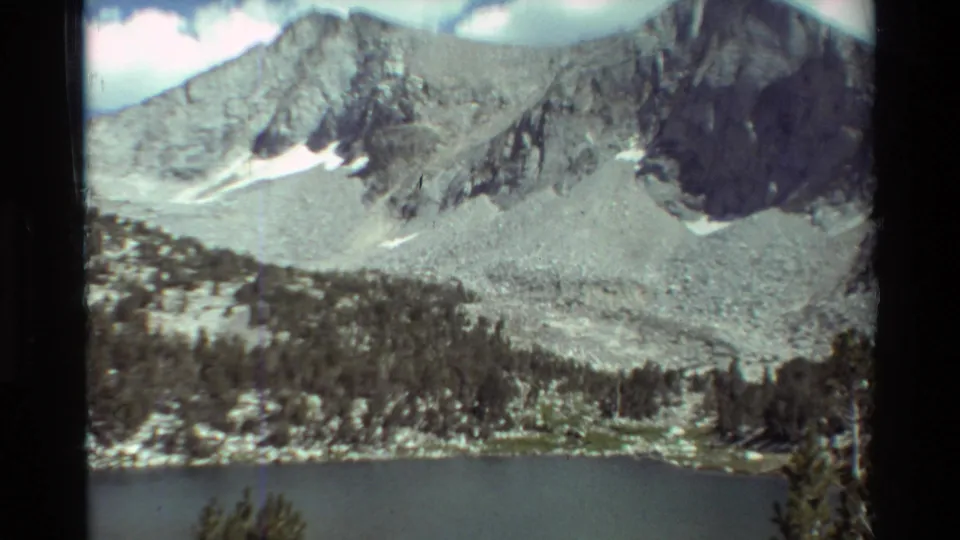 1981 8mm home-movie view of a mountain lake below rugged gray slopes, evergreen trees, clouds, and lingering snow patches.