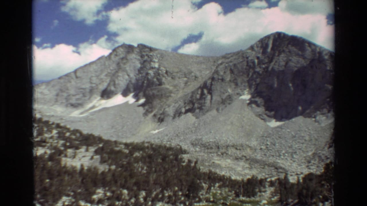 1981 8mm frame of rugged mountain slopes with snow remnants, evergreen forest at the base, and a partly cloudy sky.