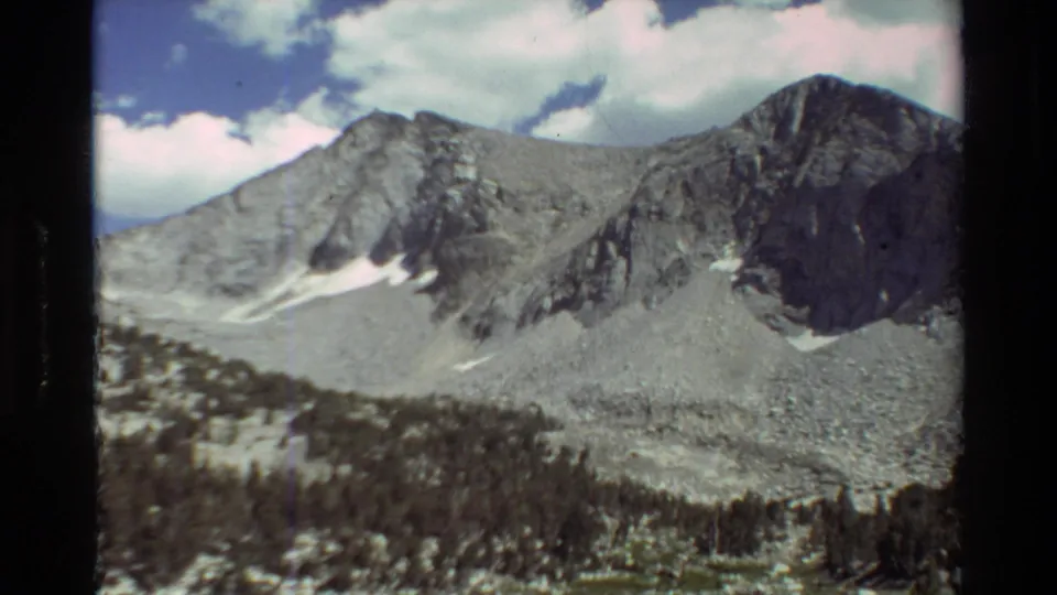 1981 8mm home-movie frame showing a closer mountain face, talus, snow patches, and dark evergreen trees at the base.