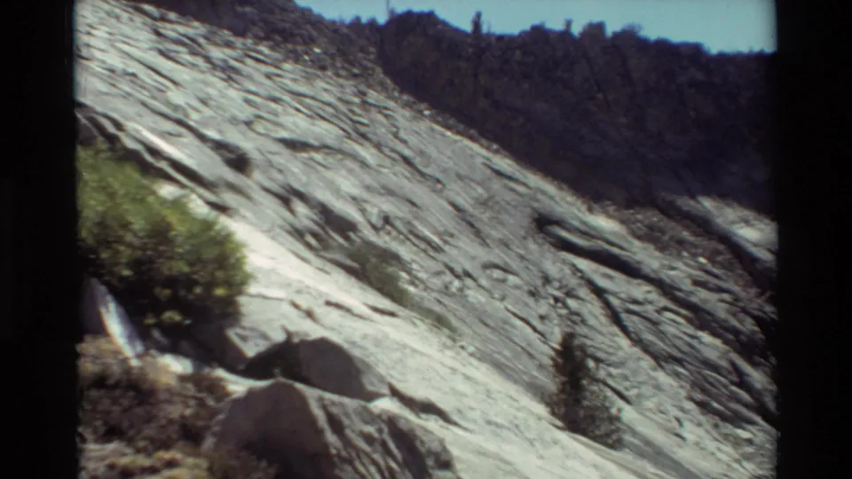 1981 8mm frame of a close rocky slope with striations, boulders, low vegetation, and a dark treeline beyond.
