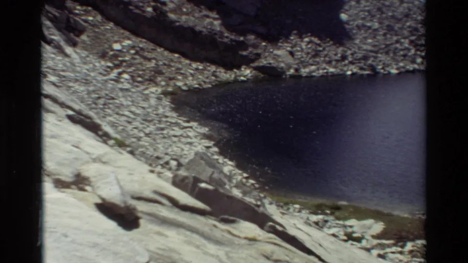 Vintage 8mm frame looking down to a dark alpine lake bordered by pale slabs, scattered stones, and sparse green growth.