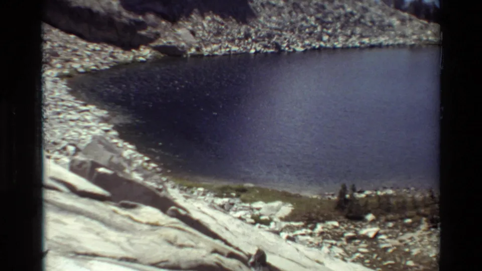 1981 8mm view of a quiet mountain lake set below scree slopes, pale rock, and a cluster of conifers near the shore.