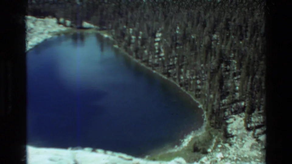 Elevated view of a blue alpine lake with forested shoreline and snow patches in 1981 British Columbia-labeled footage.