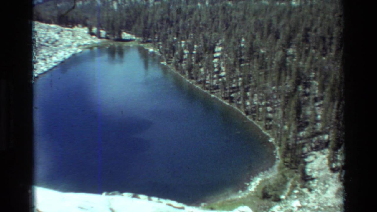 1981 8mm home-movie frame of a blue alpine lake below snowy slopes and dense conifers, with dark film edges visible.