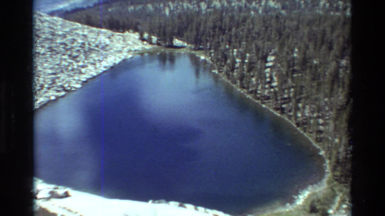 Snow-edged blue lake with pine forest and mountain terrain seen from above.