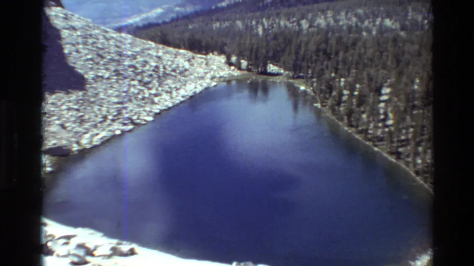 Handheld 8mm frame showing the alpine lake from above, with bright snow at the lower edge and forested slopes beyond.