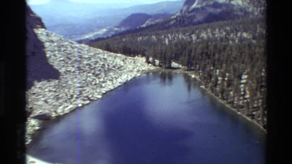 Wide 1981 home-movie frame of a blue alpine lake below snowfields, pine forest, rocky ridges, and hazy mountain distance.