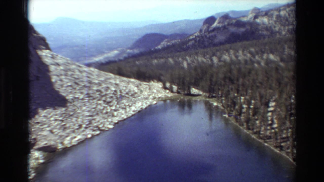 1981 8mm home-movie frame of a calm alpine lake bordered by pale rock, trees, and hazy mountain ridges.