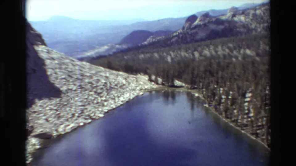 1981 8mm home-movie frame of a calm alpine lake bordered by pale rock, trees, and hazy mountain ridges.