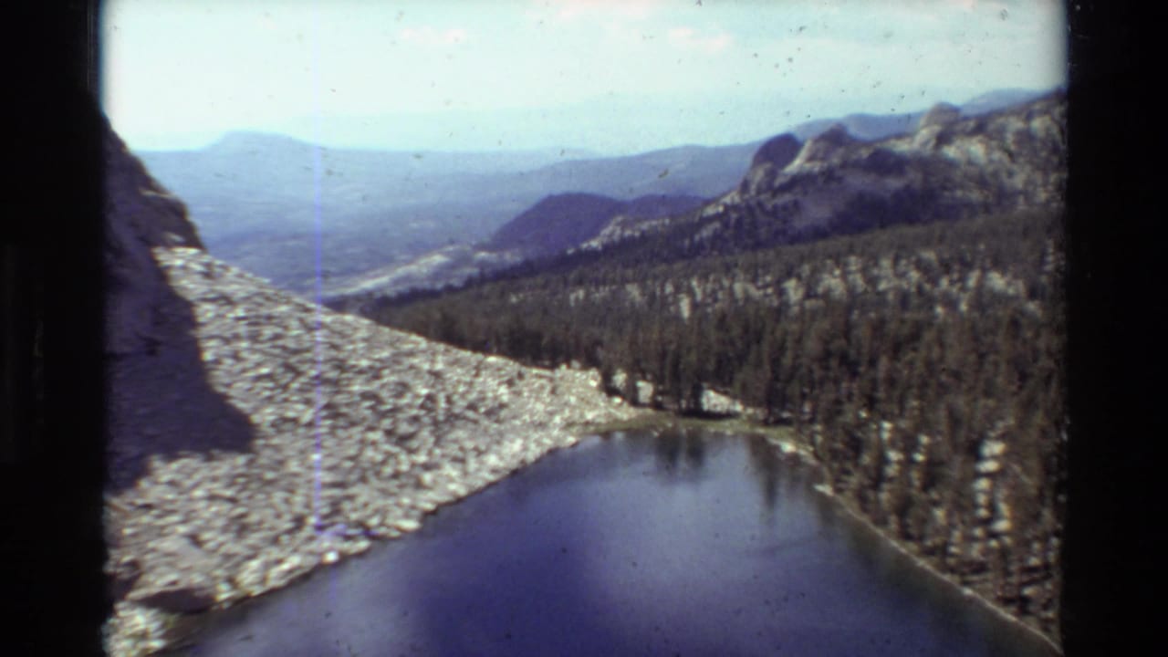 1981 8mm home-movie frame showing a still mountain lake, pale rock field, forest, and distant ridgelines.