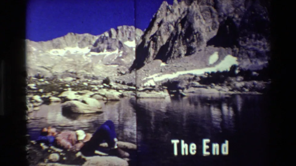 1981 8mm home-movie frame of a person in blue pants lying on a rocky lakeshore below mountain peaks and a The End title.