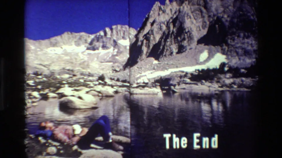 1981 8mm-style frame of a hiker resting by a mountain lake, with rocky peaks, snow patches, and a white 'The End' title.