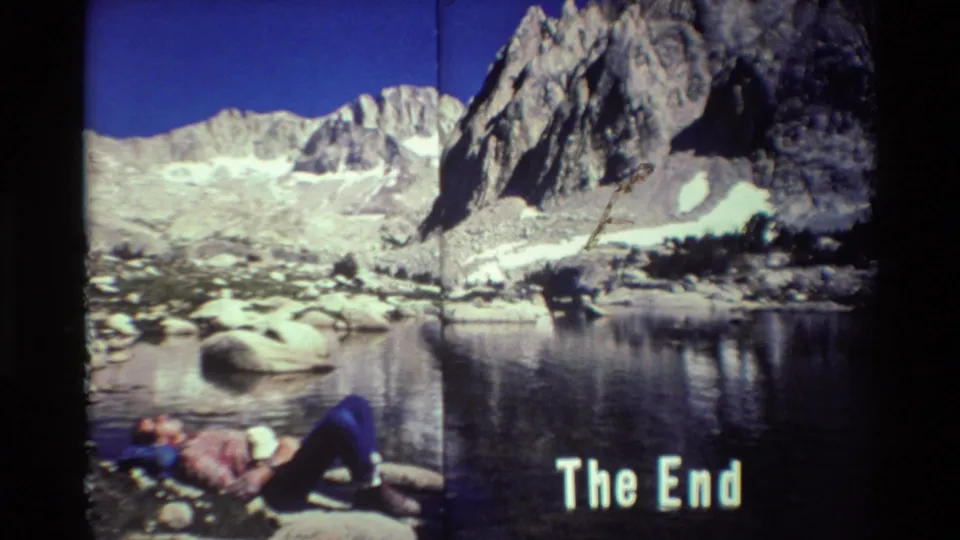 8mm home-movie frame showing a hiker on lakeside rocks below rugged mountains, with 'The End' text over the lower right.