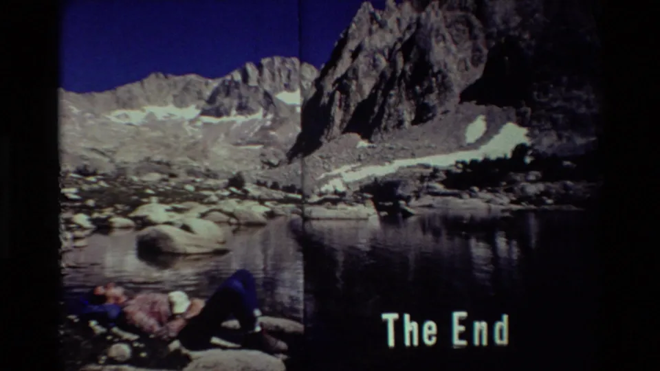 Final 8mm-style frame with a hiker resting at the rocky lakeshore, darkened frame edges, alpine peaks, and 'The End' text.