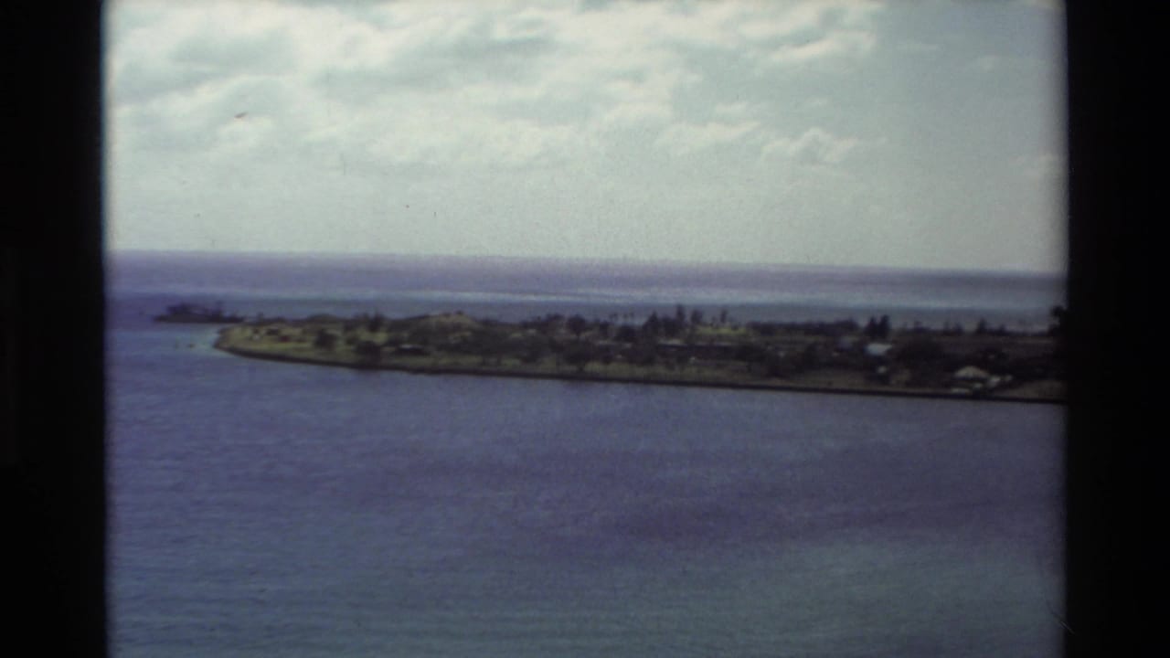 1981 Hawaii home-movie frame showing a low coastal peninsula across blue water beneath a clouded sky.