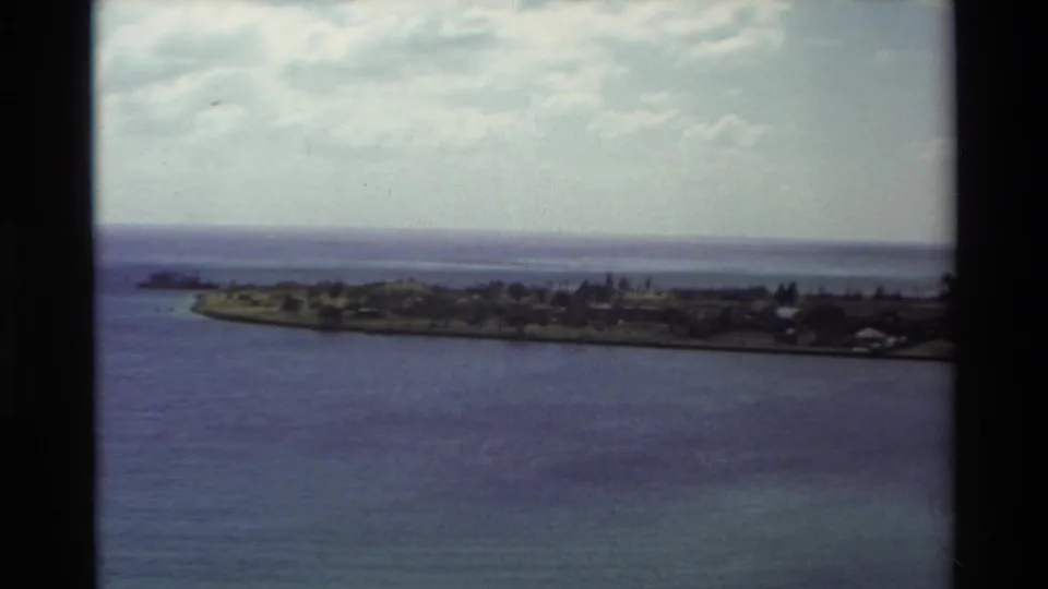 1981 Hawaii home-movie frame showing a low coastal peninsula across blue water beneath a clouded sky.