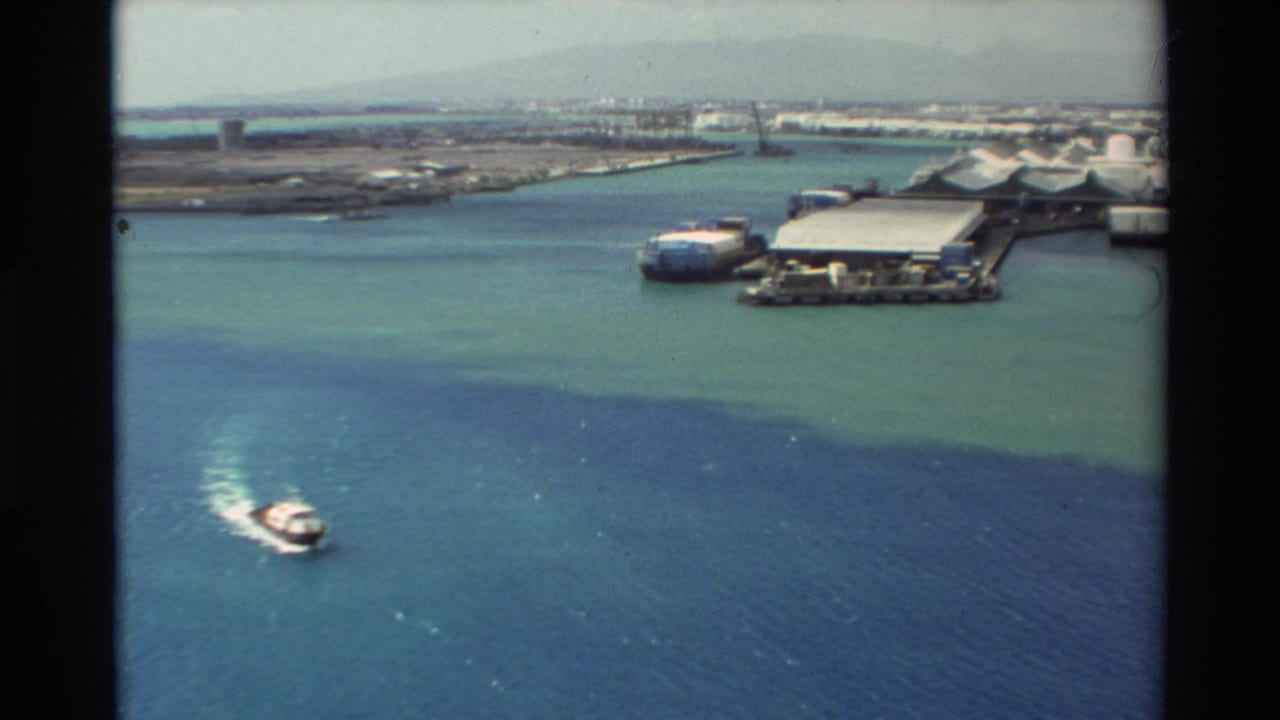 Small motorboat crossing turquoise harbor water near docks and port buildings.