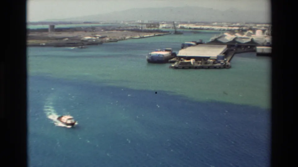 Elevated 1981 Hawaii harbor frame showing a motorboat wake, blue-green water, dock facilities, and low industrial shoreline structures.