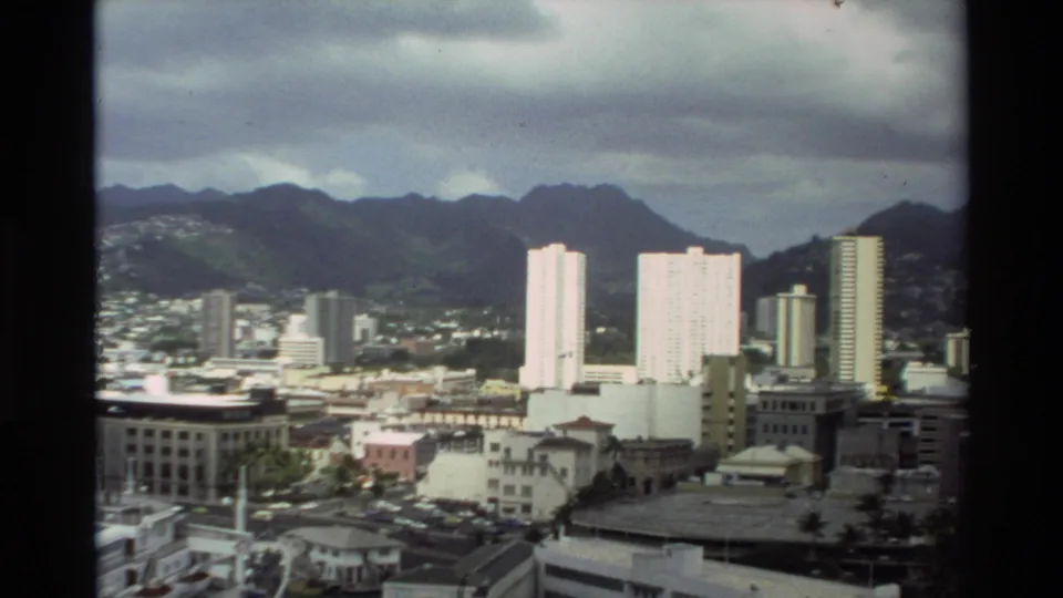 1981 Hawaii frame showing a waterfront city skyline, tall white towers, low-rise buildings, mountains, and cloudy sky.