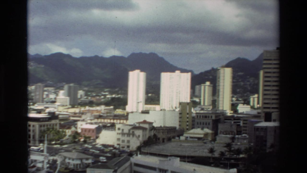 1981 Hawaii home-movie view of a city skyline with tall buildings, low-rise blocks, mountains, and overcast sky.