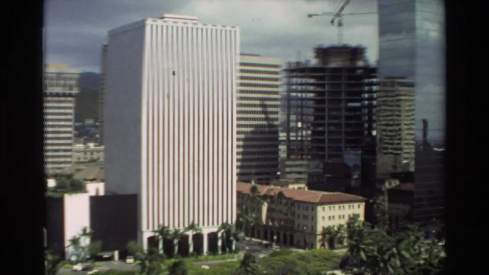 1981 Hawaii city frame with a striped high-rise, construction site, crane, nearby towers, and soft 8mm color.